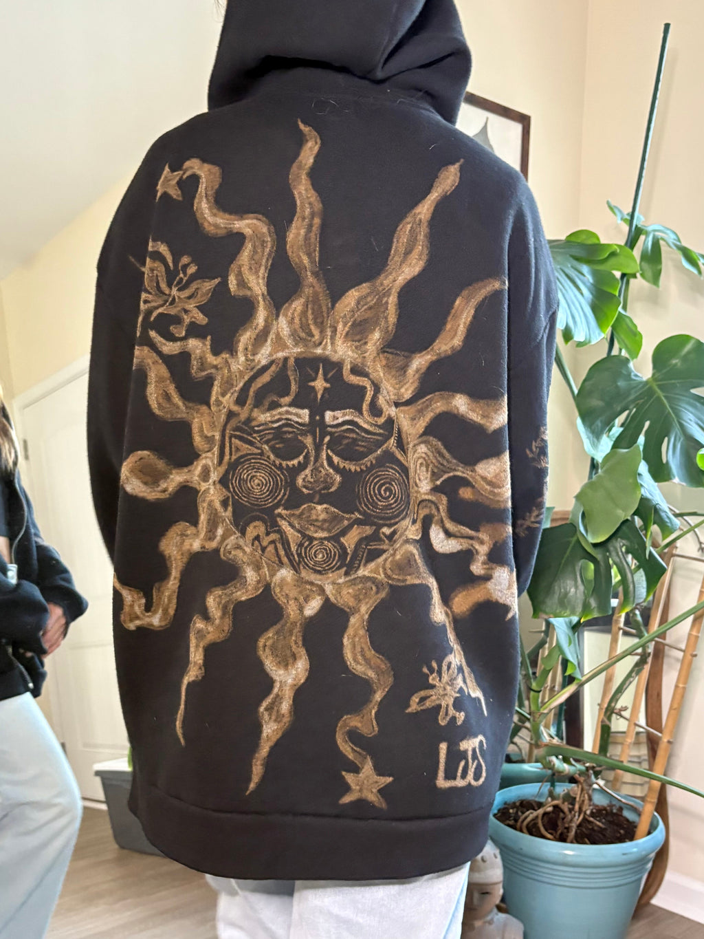 Person wearing a black hoodie with bleach art sun with butterflies design on the back in a room with plants.